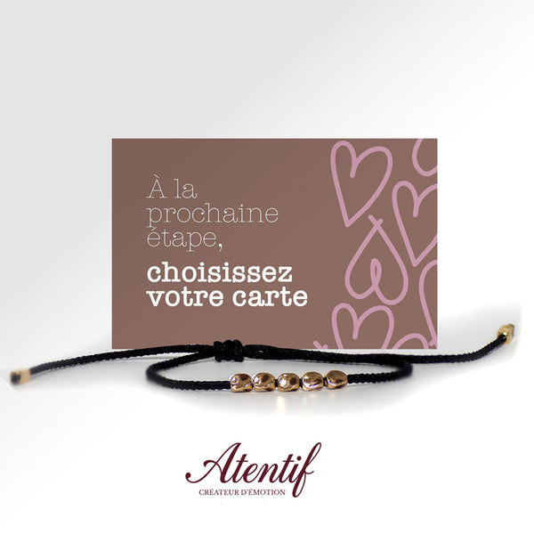 Bracelet fin en cordon noir avec cinq perles dorées. Le bracelet est posé devant une carte rose personnalisable pour un cadeau de couple.